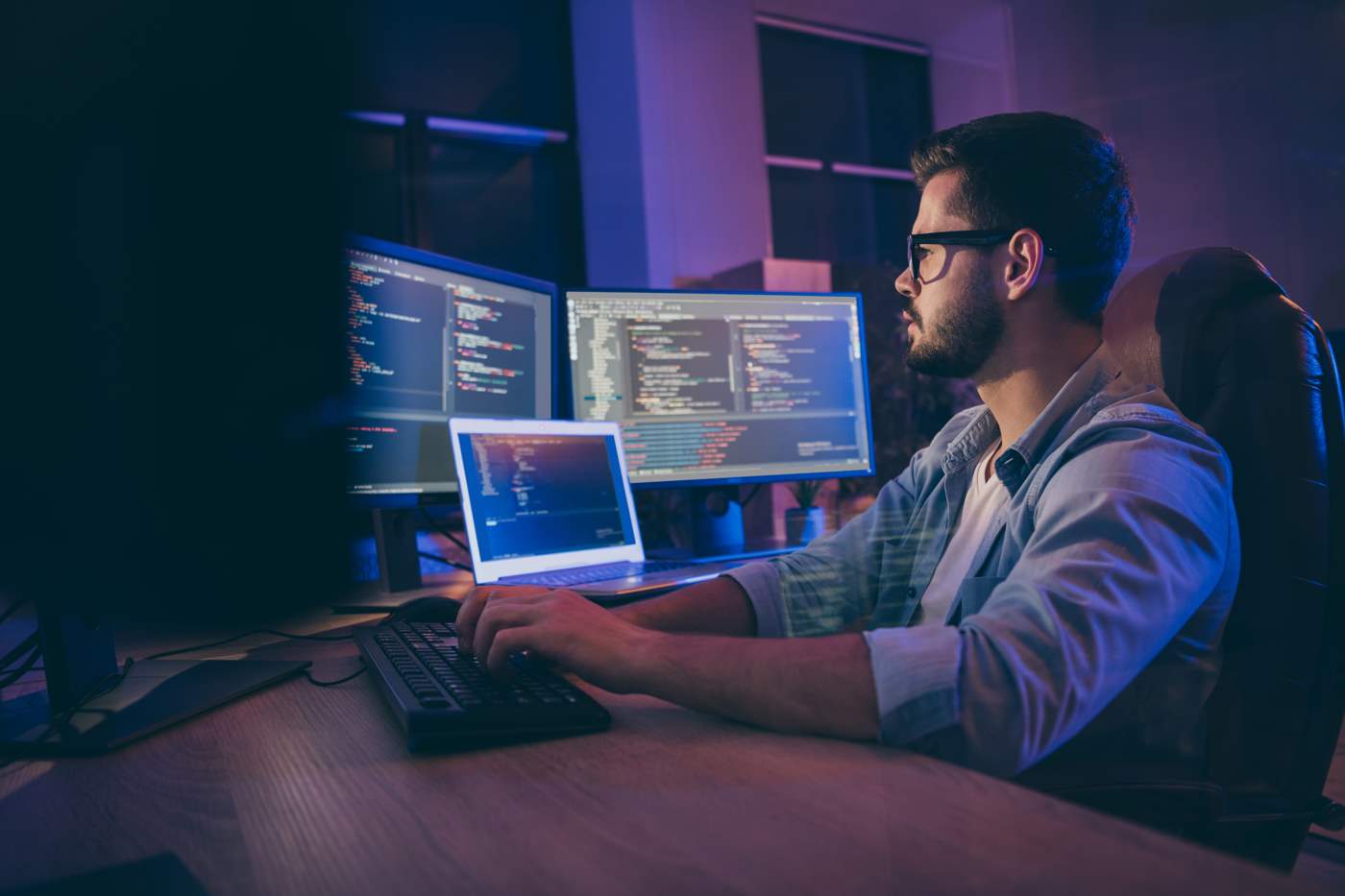Man coding in a dimly lit room with four monitors